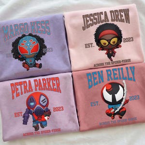 Puede incluir: Cuatro sudaderas en tonos morados y rosas, cada una con un dise&ntilde;o de personaje de dibujos animados y un nombre. Las sudaderas muestran los nombres "MARGO KESS", "JESSICA DREW", "PETRA PARKER" y "BEN REILLY", junto con el texto "EST. 2023" y "ACROSS THE SPIDER-VERSE".