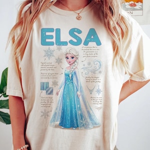 May include: Cream-colored t-shirt featuring the name "ELSA" in light blue, along with an illustration of a princess in a blue dress. The design includes snowflakes and text describing the character's features.