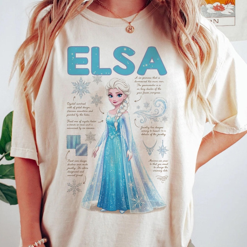 May include: Cream-colored t-shirt featuring the name "ELSA" in light blue, along with an illustration of a princess in a blue dress. The design includes snowflakes and text describing the character's features.