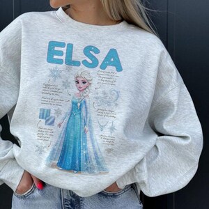 May include: A light gray sweatshirt featuring the name "ELSA" in light blue block letters. The graphic includes a cartoon image of Elsa from Frozen in a blue dress, with snowflakes and design details.