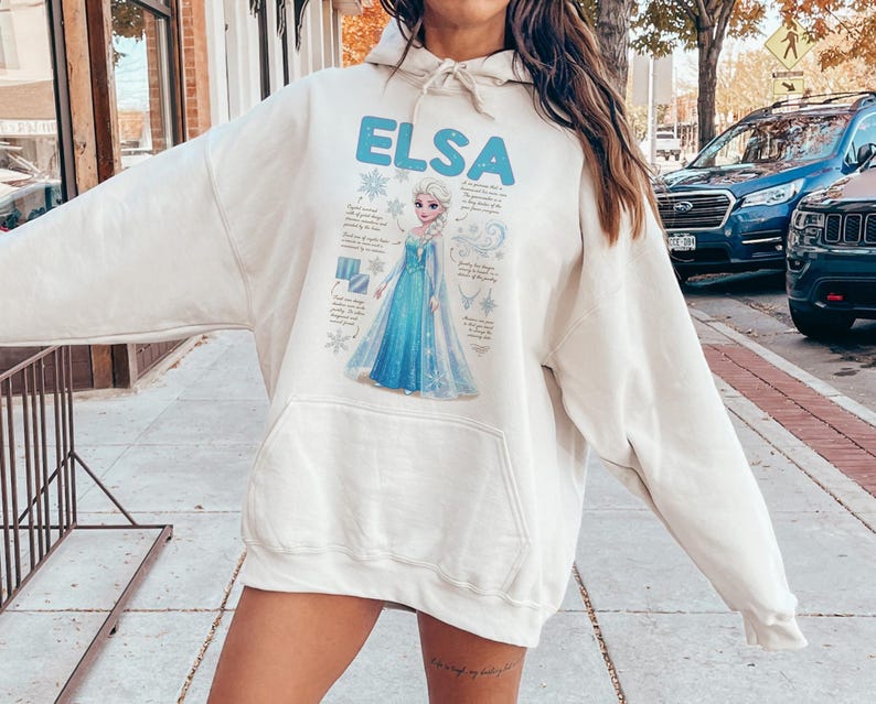 May include: A cream-colored hooded sweatshirt featuring the name "ELSA" in light blue block letters. The graphic depicts a cartoon character in a blue dress with snowflakes and text. The sweatshirt has a front pocket and a drawstring hood.