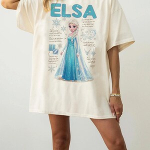 May include: Cream-colored t-shirt featuring the name "ELSA" in blue block letters above a graphic of a princess in a blue gown. The design includes snowflakes and text detailing the princess's features. The shirt is a casual, oversized fit.