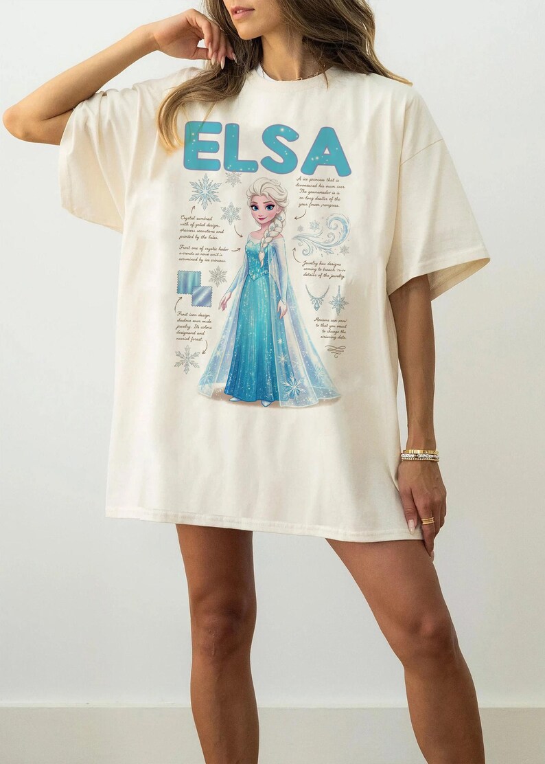 May include: Cream-colored t-shirt featuring the name "ELSA" in blue block letters above a graphic of a princess in a blue gown. The design includes snowflakes and text detailing the princess's features. The shirt is a casual, oversized fit.
