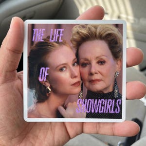 May include: A square sticker featuring a photographic portrait of two individuals with blonde hair, one wearing a gold earring and the other wearing a long, dark, beaded earring. The text THE LIFE OF SHOWGIRLS is printed in a purple glittery font.