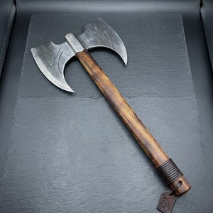 May include: A double-bit axe featuring a dark, weathered metal head with etched line patterns and a long, straight wooden handle. The handle has a dark brown leather wrap near the base, which is attached to a small, square leather tag with a raven icon.