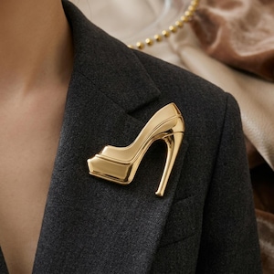 May include: A gold-colored high-heeled shoe brooch pinned to a dark gray blazer. The brooch is a stylized representation of a platform pump. The high heel is curved and slender. The brooch is a fashion accessory.
