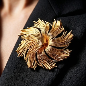 May include: A gold-colored brooch in the shape of a stylized flower, pinned to a black blazer. The brooch features numerous delicate, radiating strands, creating a textured, three-dimensional effect. The brooch is a statement piece of jewelry.