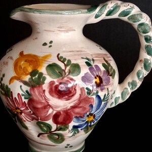 Könnte beinhalten: Eine weiße Keramikkrug mit floralem Design. Der Krug hat einen gedrehten Griff, der mit grünen Blättern verziert ist. Der Körper des Krugs ist mit roten, lila, blauen und gelben Blumen und grünen Blättern verziert.
