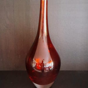 Könnte beinhalten: Eine hohe, tropfenförmige Vase in einem satten, durchscheinenden Rotton. Die Vase hat einen langen, schlanken Hals und einen abgerundeten Boden. Das Glas reflektiert die Umgebung und erzeugt einen Spiegeleffekt. Der Boden ist heller und opak.