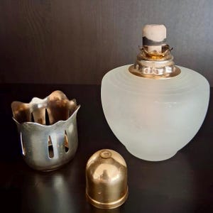 Könnte beinhalten: Eine Öllampe aus mattiertem Glas mit einem goldfarbenen Metallsockel und Dochtträger. Eine separate silberfarbene Metalltasse und eine goldfarbene Metallkappe sind ebenfalls vorhanden. Die Lampe hat eine abgerundete Form.