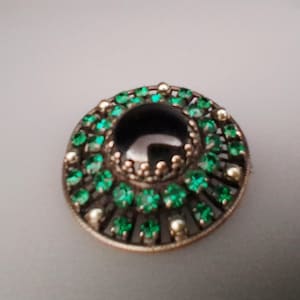 Könnte beinhalten: Eine runde Brosche mit einem großen, glatten, dunklen Mittelstein, der von zwei konzentrischen Kreisen aus kleinen, leuchtend grünen facettierten Steinen umgeben ist, gefasst in einen metallischen Rahmen mit dunkler, texturierter Oberfläche und silberfarbenen Akzenten.