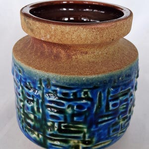 Könnte beinhalten: Eine Keramikvase mit einem strukturierten, geometrischen Design. Die Vase hat einen dunkelbraunen Innenraum, ein beiges Band in der Mitte und eine blau-grüne gemusterte Außenseite. Die Vase hat eine weite Öffnung und eine abgerundete Form.