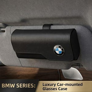 Könnte beinhalten: Ein schwarzes, rechteckiges Brillenetui für die Sonnenblende mit glatter Oberfläche und einem kreisförmigen BMW-Logo auf der Vorderklappe. Das Etui ist mit einem schwarzen Riemen an der grauen Sonnenblende befestigt und trägt den Text BMW SERIES: Luxury Car-mounted Glasses Case.
