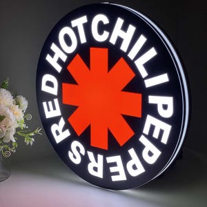 May include: A round, illuminated sign featuring the words "RED HOT CHILI PEPPERS" in white, encircling a bright red asterisk on a black background. The sign has a white border and is likely for indoor or outdoor display.