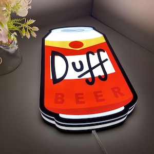 Pode incluir: Uma placa luminosa em forma de lata de cerveja Duff. A placa é vermelha com detalhes em branco e amarelo, e as palavras "Duff BEER" são exibidas de forma proeminente. Possui uma borda preta e um cabo de alimentação.