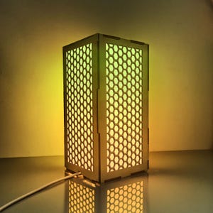 May include: A rectangular wooden lamp with a honeycomb pattern on all sides. The lamp is illuminated with a gradient of yellow and green light, and has a white power cord.