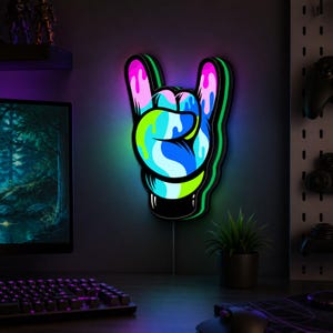 May include: A wall-mounted LED sign shaped like a hand making a rock and roll gesture. The sign features a vibrant, multi-coloured paint drip effect in shades of pink, blue, green, and white, outlined in black with a glowing neon light border.