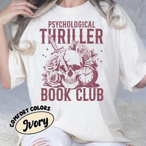 May include: An ivory t-shirt featuring a dark red graphic print of a human skull pierced by a dagger, surrounded by roses and an alarm clock. The text PSYCHOLOGICAL THRILLER BOOK CLUB is printed in a bold, serif font above and below the illustration.
