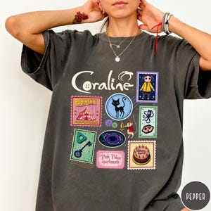 A person wearing a gray t-shirt with various colorful and whimsical images and text related to the movie "Coraline".