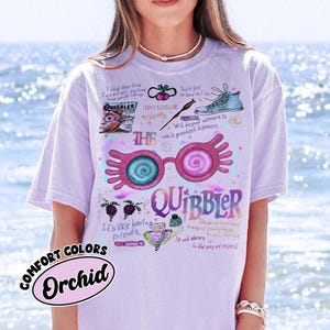 May include: Orchid-colored t-shirt featuring a whimsical design with the word "QUIBBLER" and illustrations of glasses, a shoe, and other graphics. The shirt also includes text like "Loony Lovegood" and "It's like having friends."