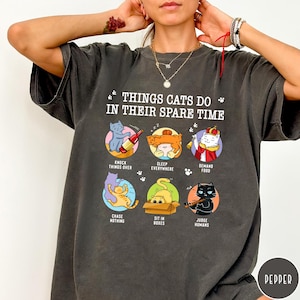 May include: A charcoal gray t-shirt featuring the text "THINGS CATS DO IN THEIR SPARE TIME" and six circular illustrations of cats performing various activities, including knocking things over, sleeping, demanding food, chasing nothing, sitting in boxes, and judging humans.