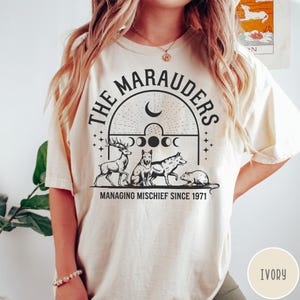 May include: An ivory short-sleeve t-shirt featuring a black graphic print. The design includes the text THE MARAUDERS and MANAGING MISCHIEF SINCE 1971, with illustrations of a stag, a wolf, a dog, and a rat beneath a crescent moon and moon phases.