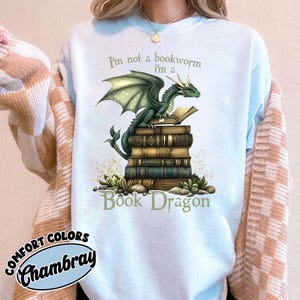 May include: A light blue crewneck sweatshirt featuring a graphic of a green dragon perched atop a stack of vintage books. The text reads, I'm not a bookworm I'm a Book Dragon. The design includes small green plants at the base of the book stack.