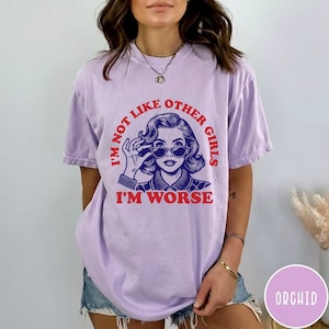 May include: A light purple short-sleeve t-shirt featuring a graphic of a person wearing cat-eye sunglasses. The red text reads I'M NOT LIKE OTHER GIRLS I'M WORSE, surrounding the blue illustration of the person's face and shoulders.