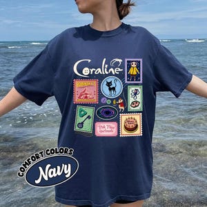 A person wearing a navy blue t-shirt with a collage of various colorful images, including a character, a cat, and other whimsical elements, against a backdrop of the ocean.