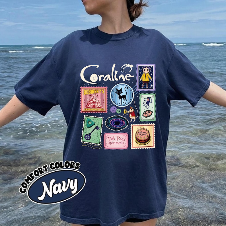 A person wearing a navy blue t-shirt with a collage of various colorful images, including a character, a cat, and other whimsical elements, against a backdrop of the ocean.
