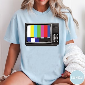 May include: A light blue t-shirt featuring a graphic print of a vintage black television set displaying a colorful SMPTE color bar test pattern on the screen. The television includes a side panel with circular knobs and a speaker grille design.