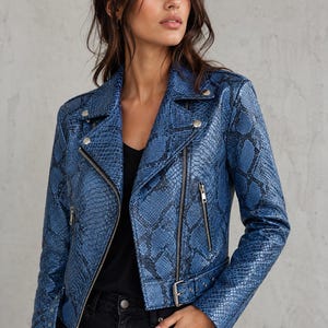 Op de afbeelding: Blauwe jas met slangenpatroon, zilveren ritsen en knopen. De jas heeft een biker-stijl met een reverskraag, asymmetrische ritssluiting en een riem in de taille. Gedragen over een zwarte top, het is een stijlvol kledingstuk.