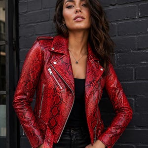 Puede incluir: Chaqueta roja con estampado de piel de serpiente, cremalleras plateadas y cinturón. La chaqueta de estilo motero tiene cuello con solapas y charreteras. Se lleva sobre un top negro y pantalones negros.