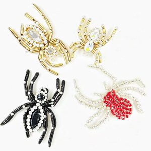 May include: Four decorative spider brooches. Two are gold with clear and white crystals, one is black with white crystals, and the last is red with white crystals. These brooches are perfect for Halloween or adding a unique touch to any outfit.