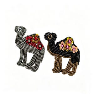 Puede incluir: Dos broches de camello con cuentas. Uno es plateado con detalles florales rojos y dorados. El otro es negro con patas doradas y detalles florales rosas y amarillos. Ambos tienen abalorios detallados sobre un fondo blanco.