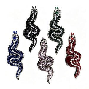 Puede incluir: Cinco broches con forma de serpiente con cuentas de varios colores: negro, verde, azul, morado y rojo. Cada broche presenta un diseño ondulado con detalles de cuentas blancas y una gema en la cabeza. Los broches están sobre un fondo blanco.