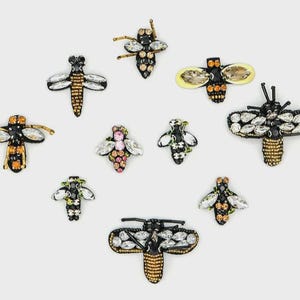 Puede incluir: Colección de broches decorativos con forma de abeja. Los broches presentan varios diseños con pedrería, cuentas y detalles metálicos. La paleta de colores incluye negro, dorado, plateado y toques de rosa y amarillo. Accesorios para añadir un toque de brillo.