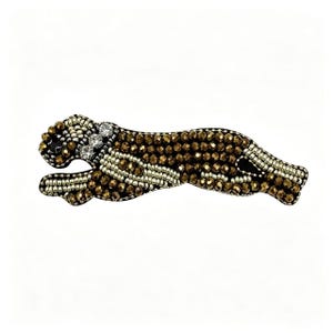 May include: A beaded cheetah brooch in a running pose. The brooch features a black base with brown and gold beads, and silver and clear crystal accents. The cheetah's body is covered in small, round, brown beads.