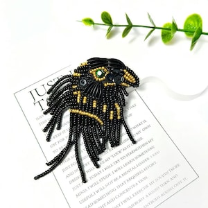 Op de afbeelding: Een geborduurde broche in de vorm van een gestileerd gezicht, met zwarte en gouden kralen en een groene edelsteen oog. De broche ligt op een wit oppervlak met een bedrukt papier en groene bladeren. De tekst op het papier luidt "JUST T".