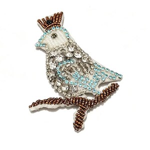May include: A beaded bird brooch with a crown. The bird is white with clear and light blue rhinestones. The crown and the branch the bird is perched on are brown. The brooch is on a white background.