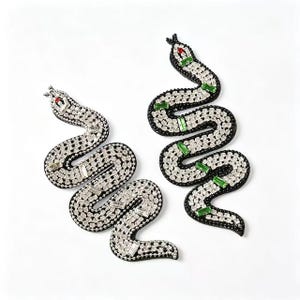 Puede incluir: Dos adornos decorativos con forma de serpiente. Las serpientes tienen un borde negro, un cuerpo de pedrería plateada y detalles rojos y verdes. Fondo blanco.