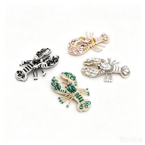 Könnte beinhalten: Vier mit Juwelen besetzte Hummer-Broschen in verschiedenen Farben. Eine ist schwarz-weiß, eine weitere rosa und gold, eine dritte silber und klar, und die letzte grün und gold. Jede Brosche ist aufwendig mit glitzernden Kristallen verziert.