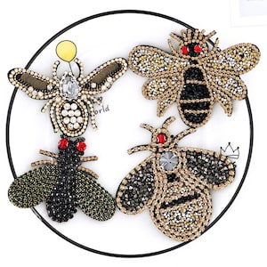 May include: Four jeweled bee brooches in various colors and designs. The brooches are embellished with rhinestones and beads, featuring red eyes and intricate wing details. One bee holds a yellow balloon.