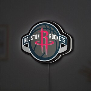 Puede incluir: Un letrero LED circular de pared con el logotipo del equipo de baloncesto Houston Rockets. El diseño incluye una letra R roja estilizada, un gráfico de balón de baloncesto gris oscuro y el texto HOUSTON ROCKETS en blanco sobre una banda azul.