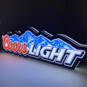 Może przedstawiać: Podświetlany znak z logo Coors Light. Znak ma czarną obwódkę z białymi i niebieskimi grafikami górskimi. Napisy "Coors LIGHT" są odpowiednio czerwone i białe.
