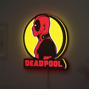 Puede incluir: Un letrero luminoso con el tema de Deadpool. El letrero presenta un gráfico rojo y negro del perfil de Deadpool dentro de un círculo amarillo, con la palabra "DEADPOOL" en letras rojas debajo. El letrero tiene un borde negro y emite un brillo amarillo cálido.