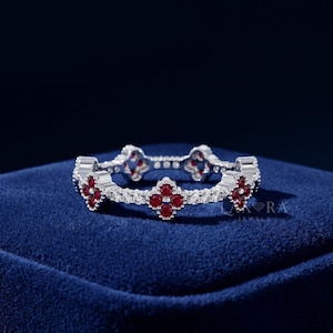 Può includere: Un anello d'argento con un motivo a trifoglio ripetuto. Ogni trifoglio è composto da quattro pietre preziose rosse, circondate da pietre preziose bianche più piccole. L'anello è posto su uno sfondo blu scuro.