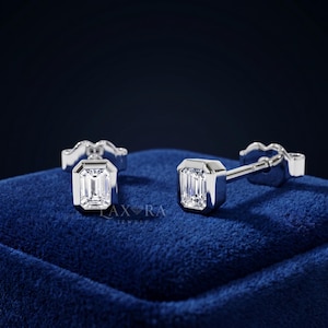 Può includere: Orecchini a lobo in argento con pietre preziose chiare taglio smeraldo. Gli orecchini sono incastonati in una montatura quadrata e sono esposti su una superficie di velluto blu. Il testo "AX RA JEWELS" è visibile.