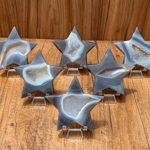 May include: Six star-shaped agate slices with banded gray and white patterns. Each piece features a central geode cavity filled with sparkling white crystals. The stars are displayed upright on small, clear acrylic stands for support.