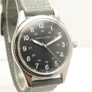 May include: A Hamilton wristwatch with a black dial, white Arabic numerals, and a 24-hour inner scale. The watch features silver-tone hands, a stainless steel case, and a textured grey fabric strap with white stitching, displaying Swiss Made text.